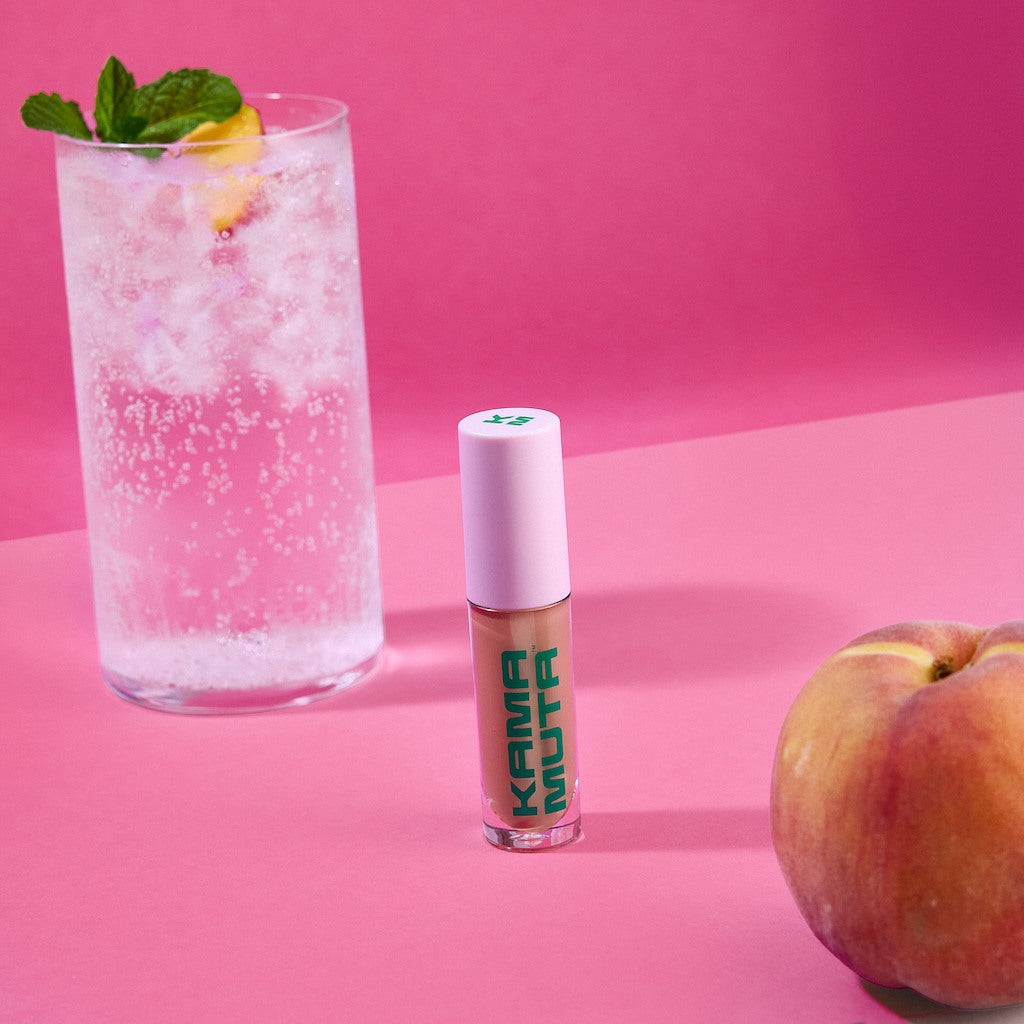 Kama Muta lip gloss with a peach and a glass of sparkling drink on a pink background