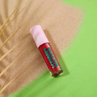 Red lip gloss with pink cap on sand against a green background